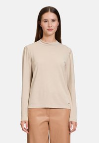 Beige Langarmshirt mit hohem Ausschnitt und gefälteltem Schulterdetail, aus einem weichen, leicht strukturierten Stoff, kombiniert mit braunen Hosen.