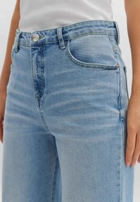 Jean en denim bleu clair avec une taille haute, une coupe droite, cinq poches, une fermeture par bouton argenté et des détails discrets de décoloration.