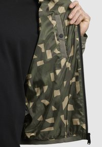Grüner Windbreaker mit gemustertem Innenfutter, das geometrische beige Formen zeigt, glatte Textur und einen Reißverschluss. Darunter ist ein schwarzes Shirt sichtbar.