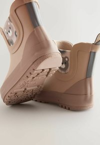 Cizme de cauciuc în bej deschis, cu suprafață texturată și un design cu un ursuleț de desene animate. Au o talpă robustă, cu model pentru aderență.