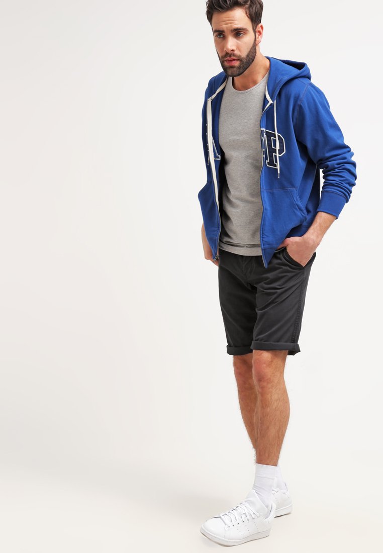 Hanorac albastru cu fermoar, cu litere albe, tricou gri dedesubt, pantaloni scurți negri și adidași albi. Material neted și croială lejera.