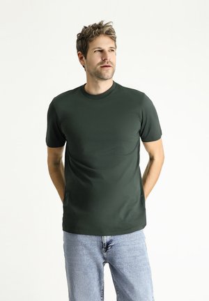 Dunkelgrünes T-Shirt mit kurzen Ärmeln aus einem glatten, elastischen Stoff und Rundhalsausschnitt, kombiniert mit hellblauen Jeans.
