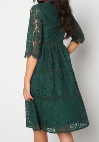 Robe en dentelle vert foncé, avec des manches 3/4, un corsage ajusté et une jupe évasée ornée de motifs floraux et une ceinture texturée.