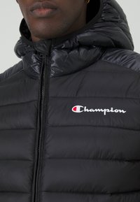 Chaqueta acolchada negra con diseño de rombos, cremallera frontal y capucha. Presenta un logo blanco de "Champion" en el lado izquierdo del pecho.