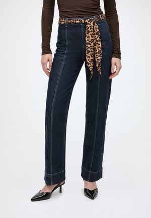 Femme portant un jean taille haute bleu foncé avec des coutures à l'avant, une ceinture léopard, un haut marron à manches longues et des talons pointus noirs.