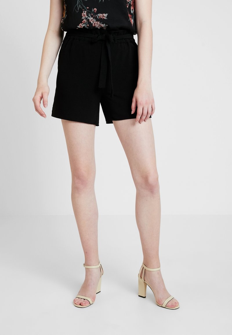 Shorts negros con cintura elástica y cinturón de lazo, con un ajuste relajado y textura suave. Combinados con sandalias de tacón alto beige.