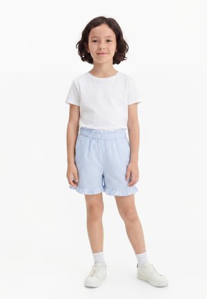 Dziecko z krótkimi ciemnymi włosami stoi uśmiechnięte, ubrane w białą koszulkę, jasnoniebieskie prążkowane spodenki z marszczonymi krawędziami, białe skarpety i białe trampki.