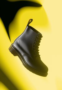 Černé kožené kotníkové boty s kulatou špičkou, texturovaným povrchem, šněrovacím designem, žlutým prošíváním a poutkem na zadní straně, umístěné na žlutém pozadí.