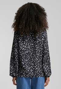 Svart bluse med blomsterlignende mønster, lange ermer og elastiske mansjetter, laget av myk stoff; løs passform; har små hvite og grå detaljer.