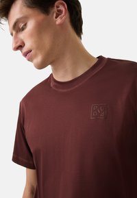 Burgundowa bawełniana koszulka z okrągłym dekoltem, krótkimi rękawami i małym wytłoczonym logo na piersi. Gładka faktura i prosty design.