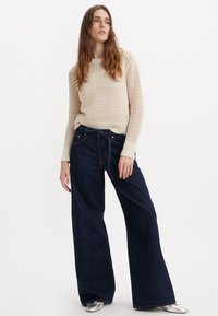 Krämfärgad stickad tröja med ett texturerat mönster och ribbade mudd, kombinerad med vidbeniga mörka denimjeans och silvriga platta skor.