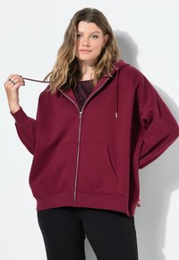 Sudadera burgundy con cremallera y un corte holgado, bolsillos delanteros, capucha con cordón y puños acanalados. Modelo llevándola sobre una blusa estampada.