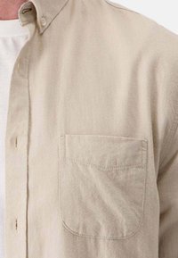 Camicia beige con bottoni, in tessuto morbido, con una tasca sul petto e colletto, caratterizzata da cuciture sottili e una vestibilità rilassata.