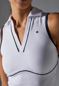 Ingrandimento di una donna che indossa un top sportivo bianco senza maniche con rifiniture blu navy e un piccolo logo nero sul petto.