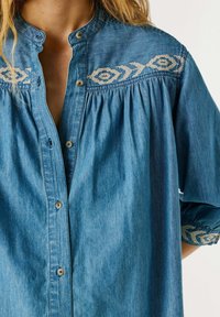 Blue denim shirt with beige geometric embroidery on shoulders and cuffs, buttoned front, worn by a person with blonde hair.
