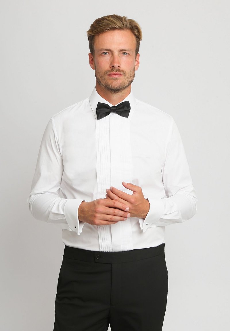 Weißes Hemd mit gefaltetem Vorderteil, schwarzer Fliege, französischen Manschetten und schlanker Passform. Tragen mit schwarzen formellen Hosen. Glatte Stofftextur.