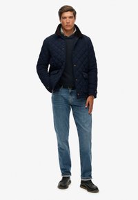 Chaqueta acolchada azul marino con cuello marrón, dos bolsillos frontales y botones dorados; combinada con unos vaqueros de color azul claro y zapatos negros, en una pose de pie.