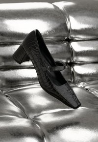 Schwarzer Lackleder-High-Heel-Schuh mit Krokodilstruktur, quadratischer Spitze und Knöchelriemen, vor einem metallisch silbernen Hintergrund.