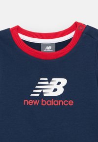 Mørkeblå New Balance skjorte med rød og hvid kant rundt om halsen og hvidt logo over rødt "new balance" tekst på brystet, snapknap på skulderen.