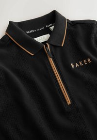 Sort polo t-shirt med et tekstureret mønster, gyldne accenter på kraven og lynlåsen, med "BAKER" broderet i guld på forsiden.