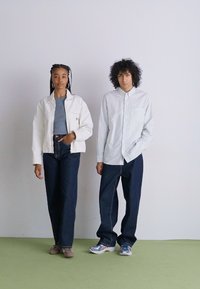 Hvit jakke over grå topp, blå høyt liv jeans; stripete skjorte med knapper, mørke jeans; begge har på seg uformelle sko; glatte teksturer.