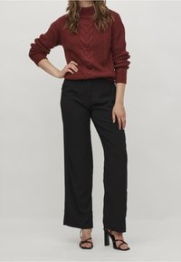 Nainen, jolla on päällä marooninvärinen palmikkoneule, mustat leveälahkeiset housut ja mustat säänmukaiset korkokengät, seisoo käsivarsi lantiolla.