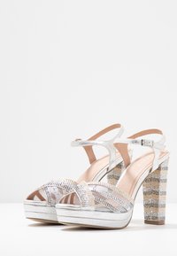 Stříbrné platformové sandály s texturovanými pásky, otevřenou špičkou a páskem kolem kotníku. Blokový podpatek má pruhovaný vzor. Hladký, lesklý materiál.