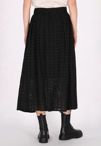 Černá maxi sukně s vzorem broderie anglaise a květinovými výřezy, s vysokým pasem a volným střihem. V kombinaci s černými kotníkovými botami.