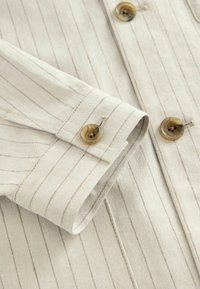 Detaliile cămășii bej cu dungi, incluzând o mansetă pliată cu un buton mare și rotund, de culoare maro. Materialul are o textură netedă și o strălucire subtilă.