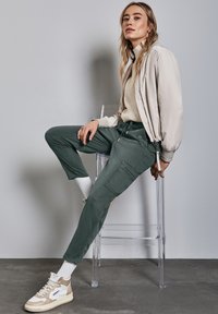 Frau trägt eine beige Bomberjacke, einen cremefarbenen Pullover, grüne Cargohosen, weiße Socken und beige Turnschuhe, sitzt auf einem klaren Acrylhocker.