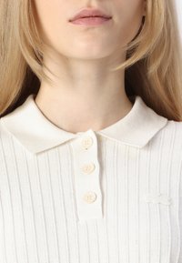 Polo de color crema con un diseño texturizado de canalé, un cuello y cuatro botones crema en la tapeta. Pequeño logo a la izquierda.