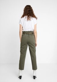 Olivově zelené kalhoty s vysokým pasem, opaskem v pase, zúženými nohavicemi a dvěma zadními kapsami, spárované s bílým tričkem a botami na podpatku.