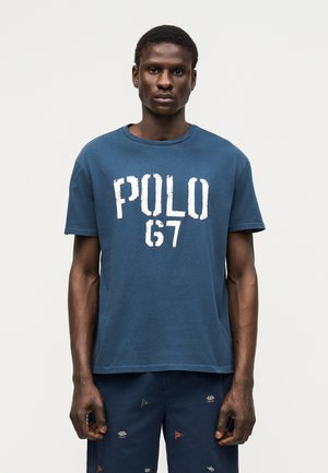 Mężczyzna ubrany w niebieską koszulkę "POLO 67" oraz granatowe szorty z małymi haftowanymi flagami, stojący na tle jednolitego jasnego tła.