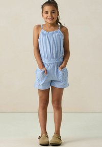 Rompers rayés bleus avec des fines bretelles, un décolleté froncé, une taille élastique et des poches en forme de cœur ; porté avec des chaussures marron clair.