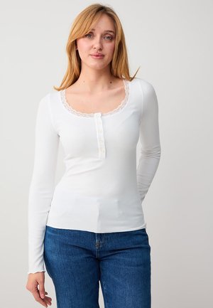 Junge Frau mit blondem Haar, die ein weißes Langarmoberteil mit Knöpfen und Spitzensaum trägt, kombiniert mit blauer Jeans, vor einem schlichten Hintergrund stehend.