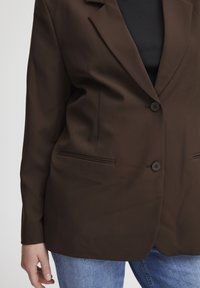 Blazer marron avec une coupe ajustée, deux boutons et poches avant. Le tissu semble lisse avec un léger éclat ; associé à un haut noir.