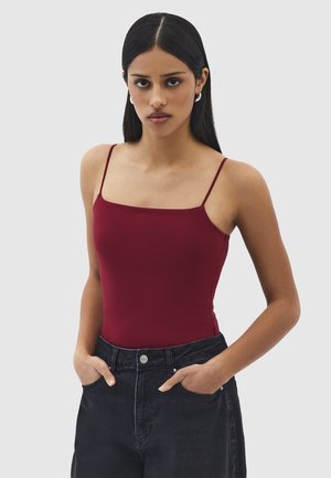 Donna con capelli lunghi e scuri che indossa orecchini a cerchio d'argento, un top aderente bordeaux con spalline sottili e jeans neri ad alta vita con le mani in tasca.