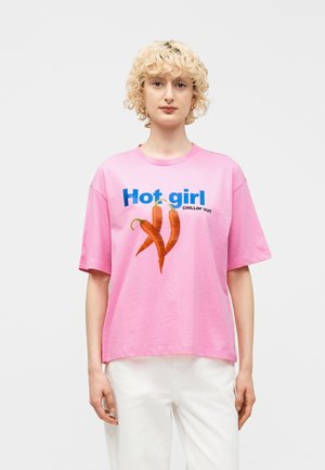 Młoda osoba o kręconych blond włosach, ubrana w luźną różową koszulkę z papryczkami chili i napisem „Hot girl chillin' out”, białe spodnie, neutralne tło.