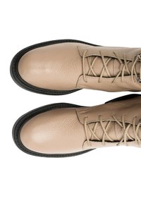 Bottes beiges en cuir à lacets avec bouts arrondis, semelles en caoutchouc noir et œillets en métal, vues de dessus sur un fond blanc.
