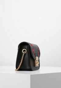 Musta nahkalaukku punaisella kukkaompelulla, kultaketjuolkaimet ja kultalukko, joka lepäilee valkoisella pedestaalilla tasaisella taustalla.