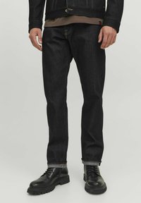 Jean en denim foncé avec une coupe droite, des coutures jaunes et des ourlets retroussés. Associés à des bottes noires à lacets et un haut marron sous une veste.