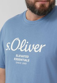 Blauwe katoenen t-shirt met een ronde hals en korte mouwen, voorzien van witte tekst die luidt: "s.Oliver ELEVATED ESSENTIALS SINCE 1969."