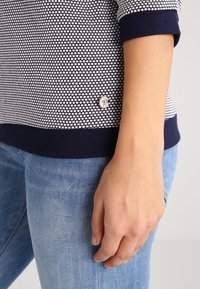 Marinblå och vit mönstrad stickad topp med ribbade manschetter och en silverfärgad logoknapp på nedre sidan, bärs tillsammans med ljusa denimjeans.