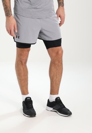 Pantalones cortos deportivos grises con capa de compresión negra debajo, hechos de tejido ligero, cuentan con una cinturilla elástica y un logotipo en el lateral. Zapatillas de correr negras completan el atuendo.