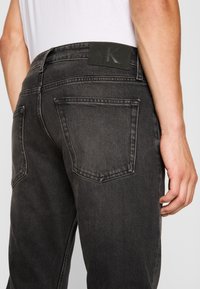 Jeans de mezclilla negra con una sutil textura desgastada, diseño de cinco bolsillos y una etiqueta rectangular de cuero negro en la parte trasera de la cinturilla.