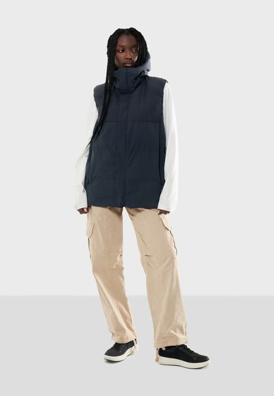 Marinblå puffer-väst med hög krage, framfickor och slät textur, kombinerad med krämfärgade cargobyxor och svarta sneakers.