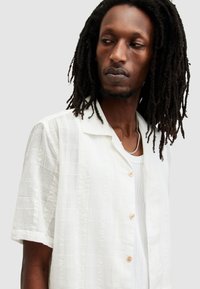 Camisa blanca de manga corta hecha de una tela texturizada con un diseño estampado, que presenta un cierre de botones en la parte delantera y un cuello relajado.