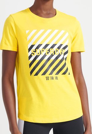 Camiseta de algodón amarilla con mangas cortas, que presenta un diseño gráfico de rayas diagonales en blanco y negro y la palabra "SUPERDAY" en letras negritas.