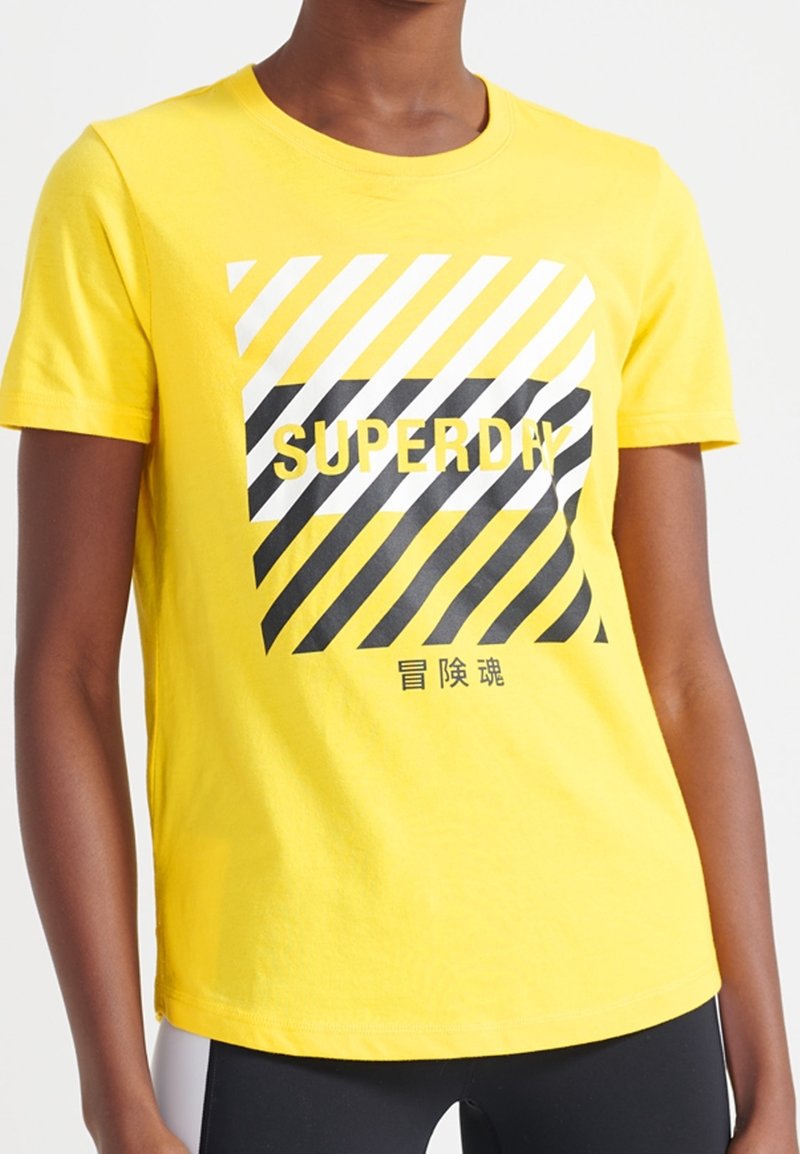 Camiseta de algodón amarilla con mangas cortas, que presenta un diseño gráfico de rayas diagonales en blanco y negro y la palabra "SUPERDAY" en letras negritas.