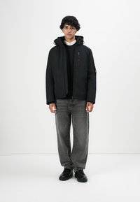 Ung mand iført en sort hættejakke, sort sweater, hvid skjorte, bleget grå jeans og sorte sko, stående mod en hvid baggrund.